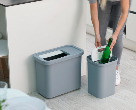 A lady disposing a glass bottle into one of the trash bin compartment. The other compartment is separated and sitting beside a kitchen counter.