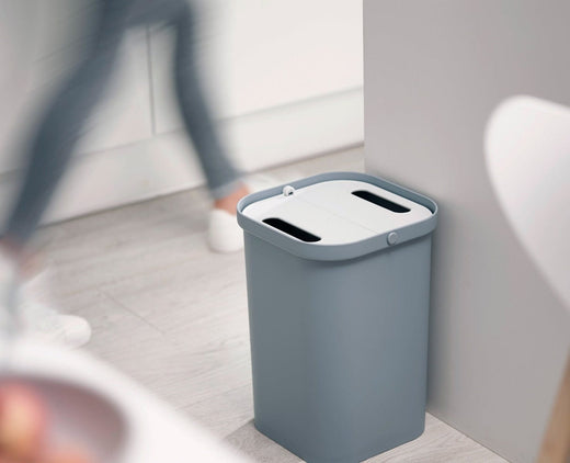 A recycling caddy with a grey base and a closed white lid is situated next to the kitchen counter.